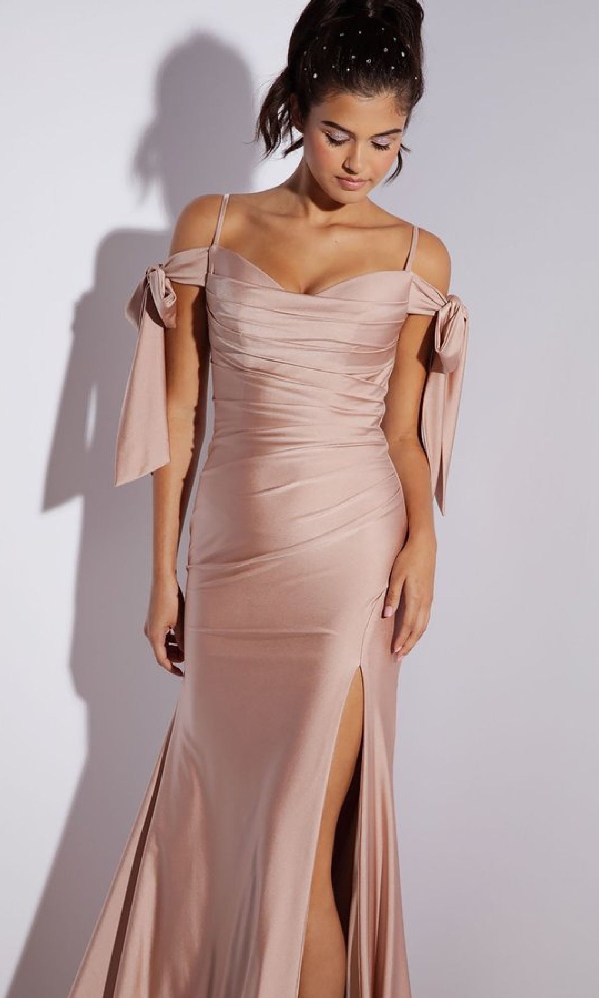 Woman in the Shoulder-Ties Long Jersey Prom Dress: Eureka 9181—a light pink, off-the-shoulder gown with side slit and bow sleeve details. This affordable evening dress is perfect for making a statement against a plain background.